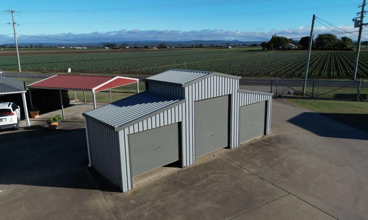 Grey Oz barn-style shed beside a red carport with crop rows behind